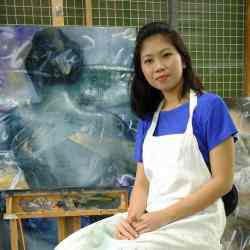 Photo of Rina Lee Garibay (Filipina sitting in front of a painting, wearing a white apron over a blue shirt)